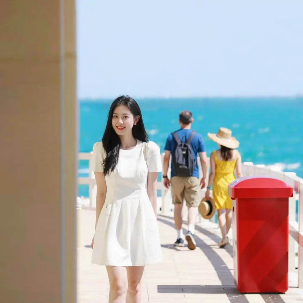 Woman in a white dress on a seaside boardwalk, with two tourists and a red bin behind her